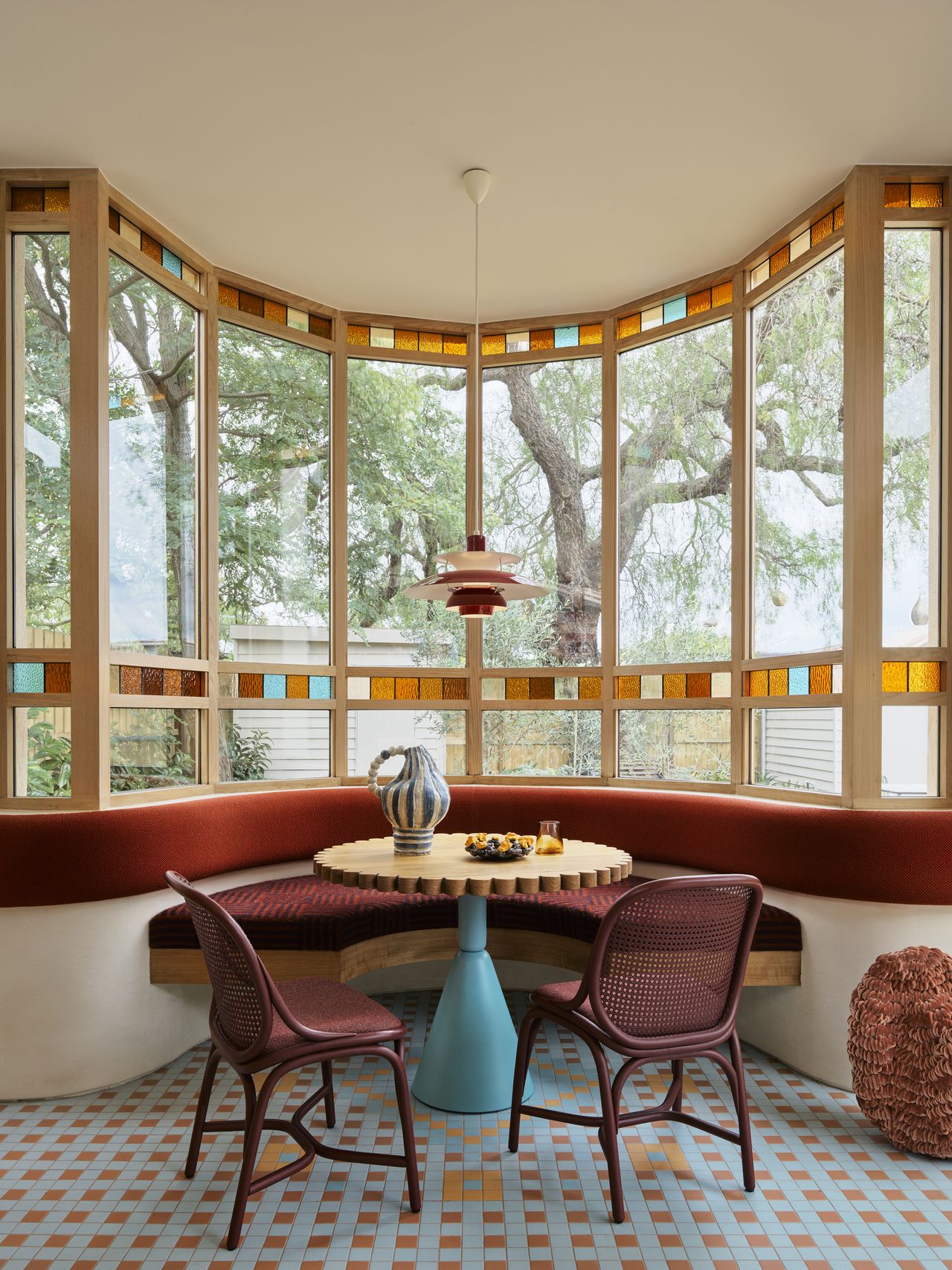 Image of an off-white dining nook with oxblood red upholstery and seating. The wall is made of windows with a stained glass trim.