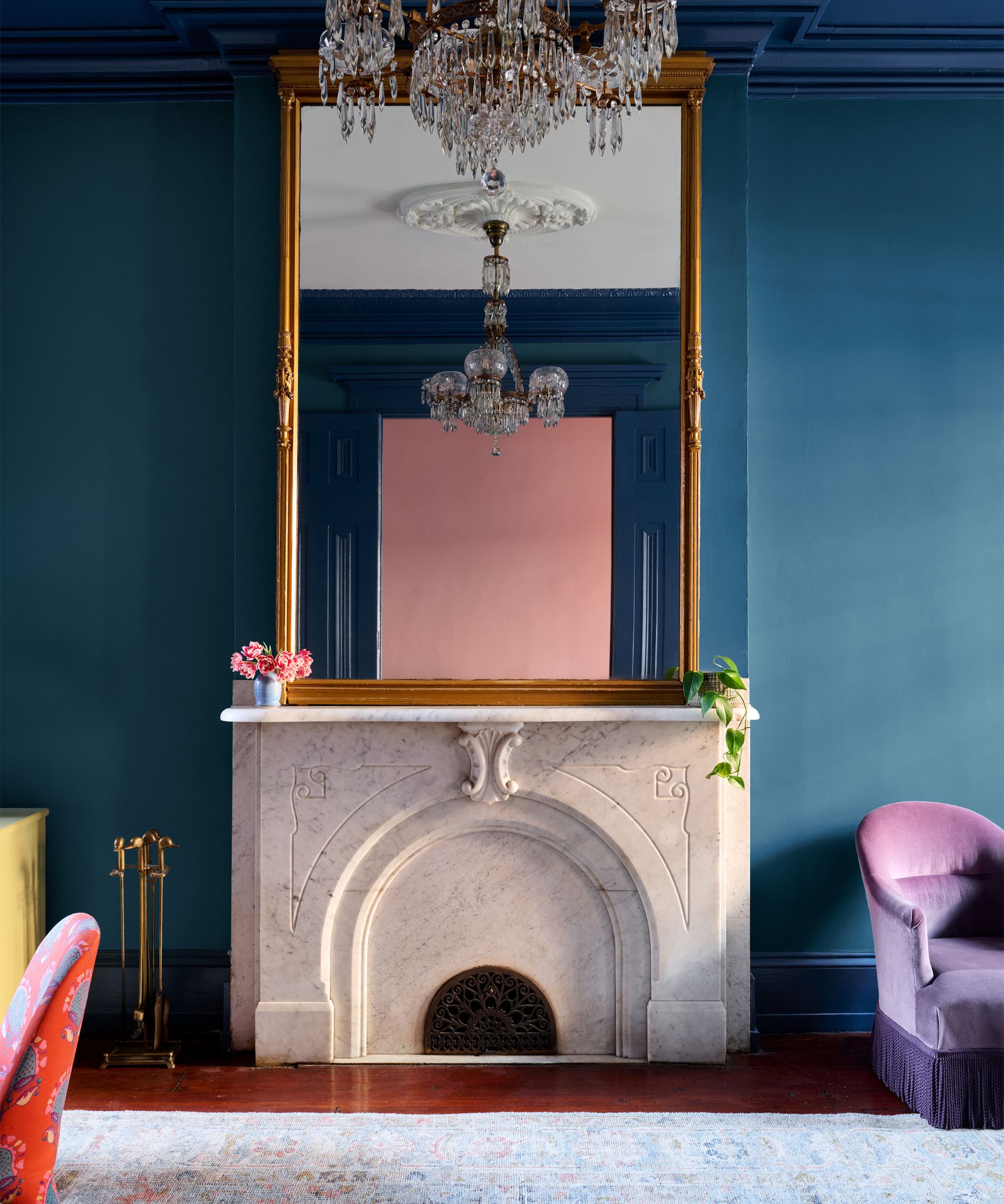 White marble fireplace against a teal wall, featuring a large gold mirror reflecting a pink wall and crystal chandelier.