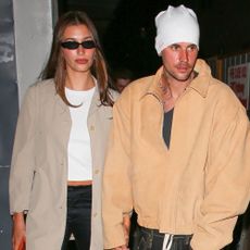 Hailey Bieber holds hands with Justin Bieber wearing a beige chore jacket with satin trousers and kitten heel flip flops.