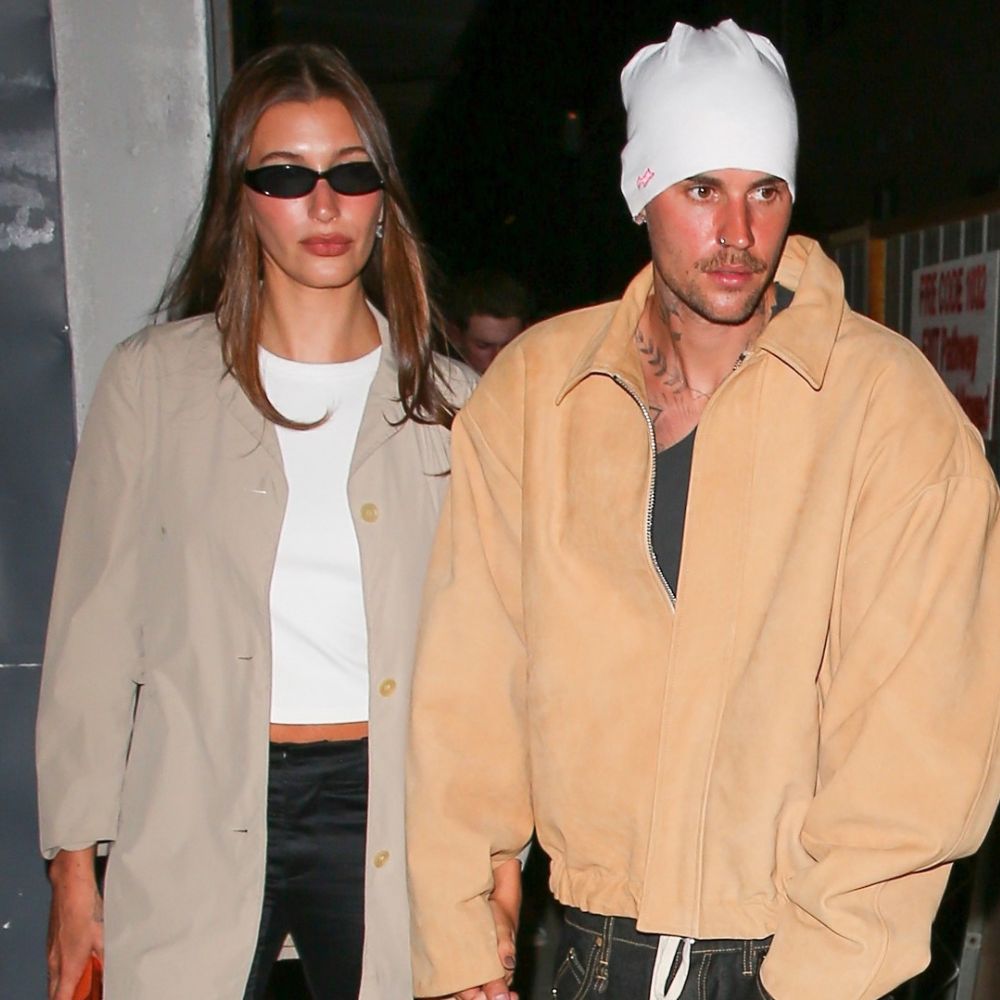 Hailey Bieber holds hands with Justin Bieber wearing a beige chore jacket with satin trousers and kitten heel flip flops.