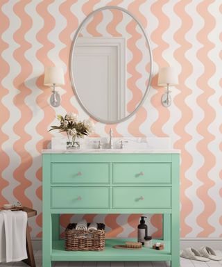 A bathroom with with and peach wavy painted walls, a silver round mirror, two silver wall sconces, and a mint green vanity unit with four drawers and a basket underneath