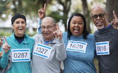 A multiracial group of four adults, two couples, having fun at a race competition. They are standing side by side wearing numbered sports bibs, smiling and looking at the camera, making the number one finger sign.