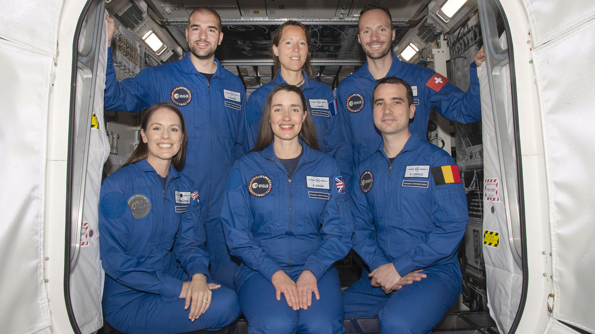 six people in blue flight suits pose in the doorway of a simulated space module