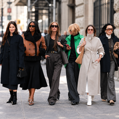 Four women in layered fall outfits, including sweaters, skirts, jeans, scarves, and loafers, walk along a city street.