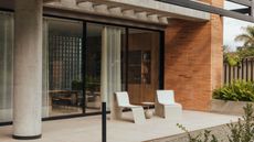 A small patio with two chairs and a tale in the middle. There are plants on the right side of the image growing from a plant bed. 