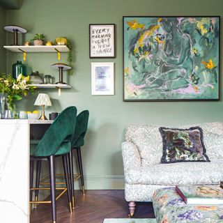 a green open plan kitchen/living area with a breakfast bar and velvet upholstered bar stools, fabric covered sofa and striking art work on the wall