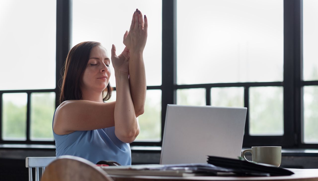 Woman sitting at a table with an open laptop in front of her. She has her eyes closed and her arms are intertwined in eagle pose. She is wearing a light blue sleeveless top and has shoulder-length brown hair