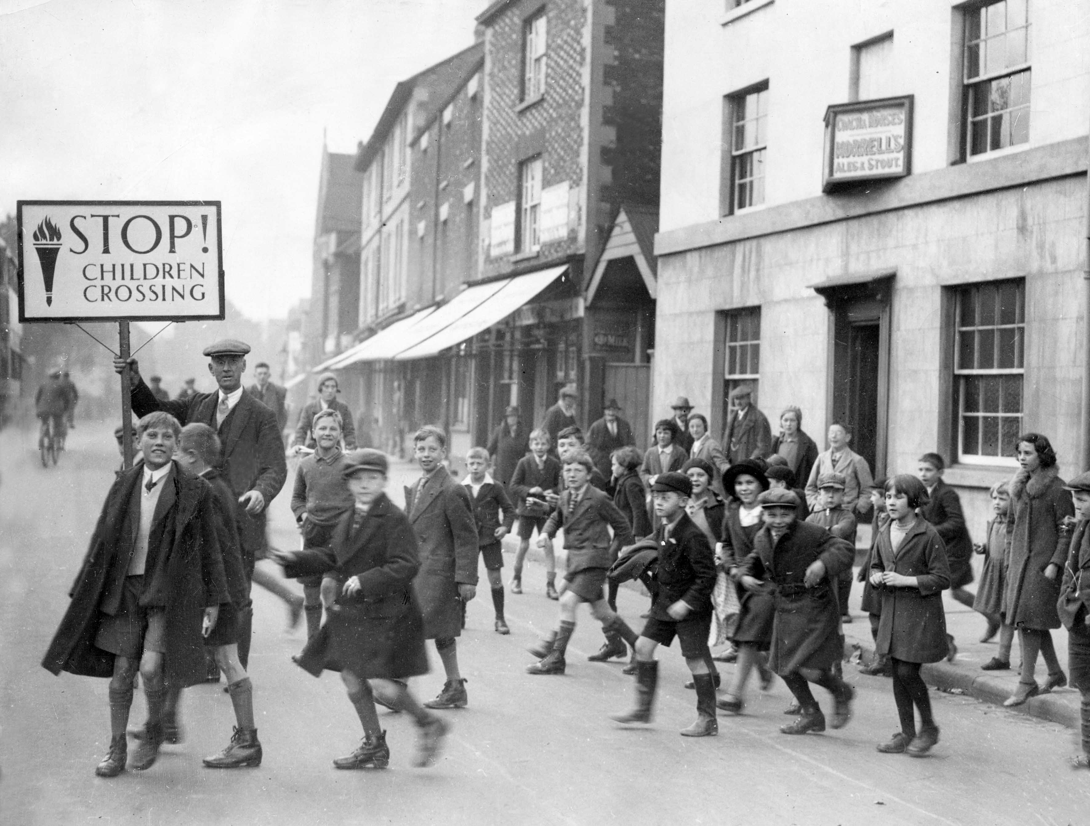 A man in a flat cap holds up a "Stop! Children Crossing" sign as a crowd of schoolchildren in coats and caps run and walk across a street in Oxford.