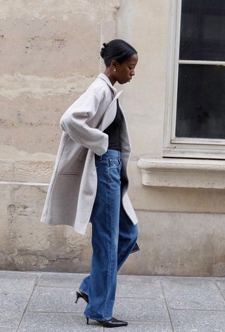 The winter capsule wardrobe item, baggy jeans, is shown in a photo of a woman walking down the sidewalk wearing a gray wool coat over a black sweater styled with baggy jeans layered over black tights styled with black mules