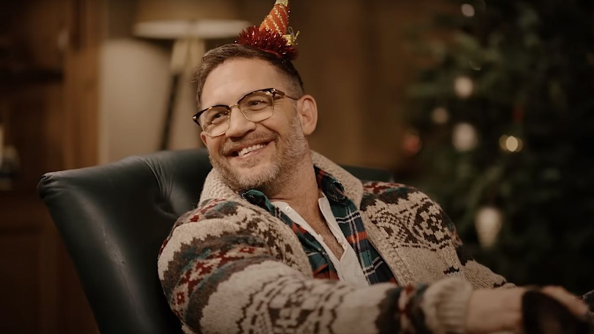 Tom Hardy smiling wearing a red party hat and a Christmas sweater in commercial for Jo Malone London.