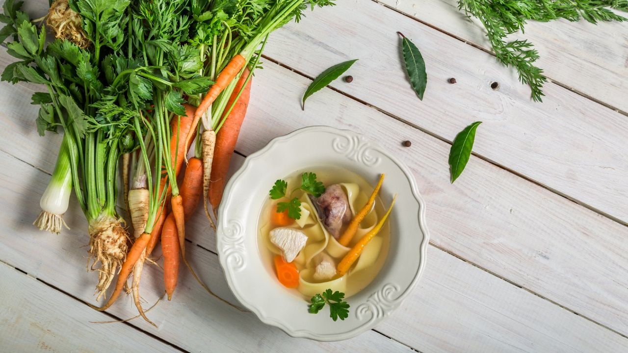 Carrots, celery root, and leeks next to a bowl of vegetable noodle soup