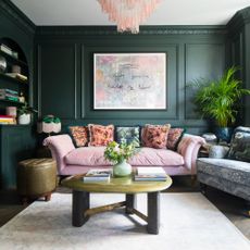 A dark green-painted living room with a pink velvet sofa with five floral-print velvet cushions with a fringed trim