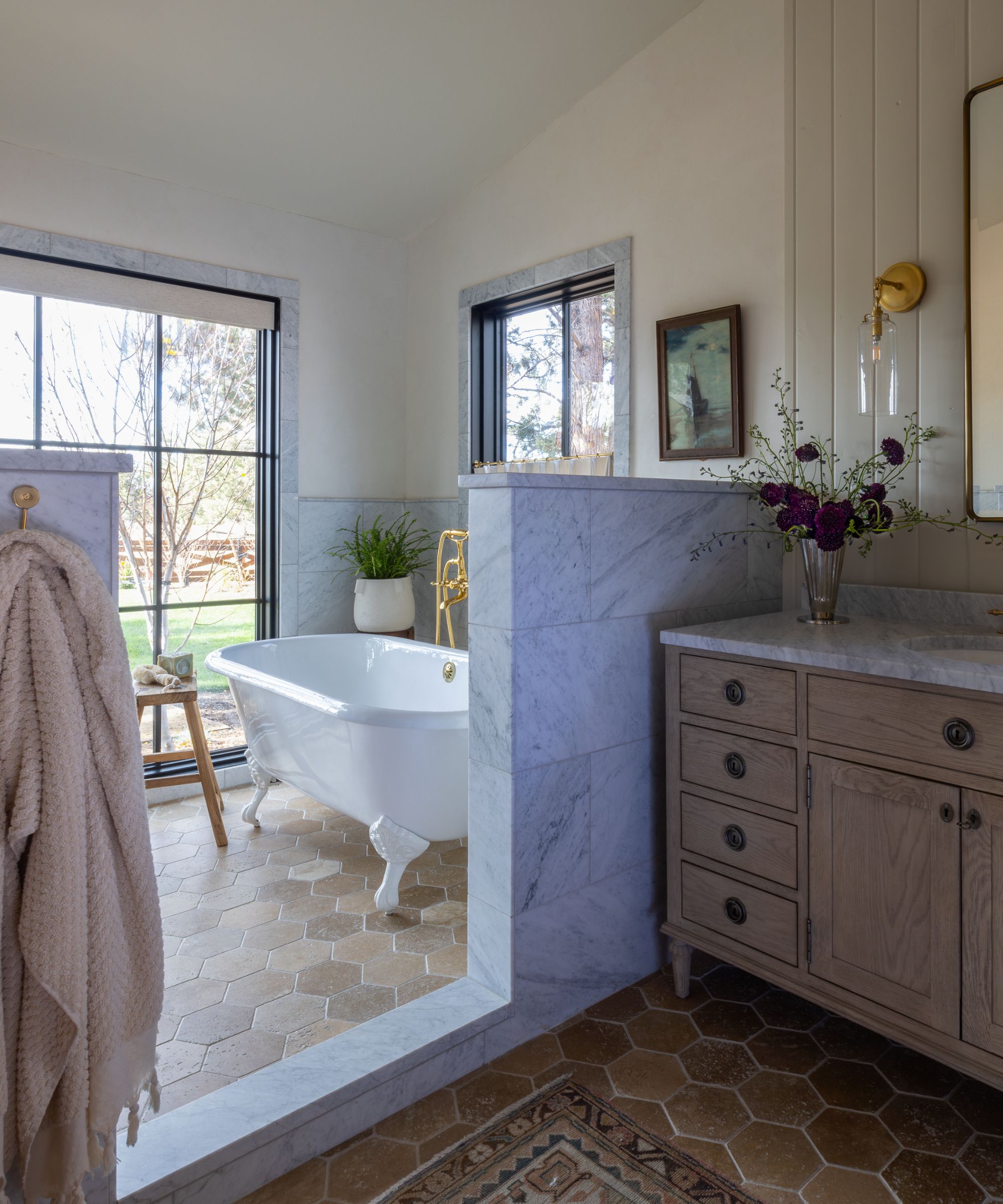 A large bathroom with a freestanding white clawfoot tub, marble half-wall divider, and hexagonal terracotta floor tiles. On the other side of the divider is a wooden vanity with a marble top and brass fixtures.