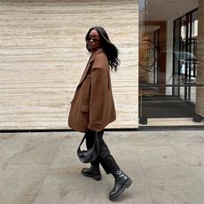 influencer wears brown coat, black pants, and chunky soled boots while carrying a black shoulder bag