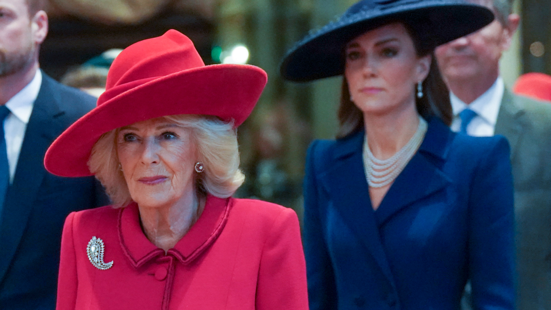 Queen Camilla wears a red coat dress and matching hat while Princess Kate stands behind her wearing a navy coat and hat