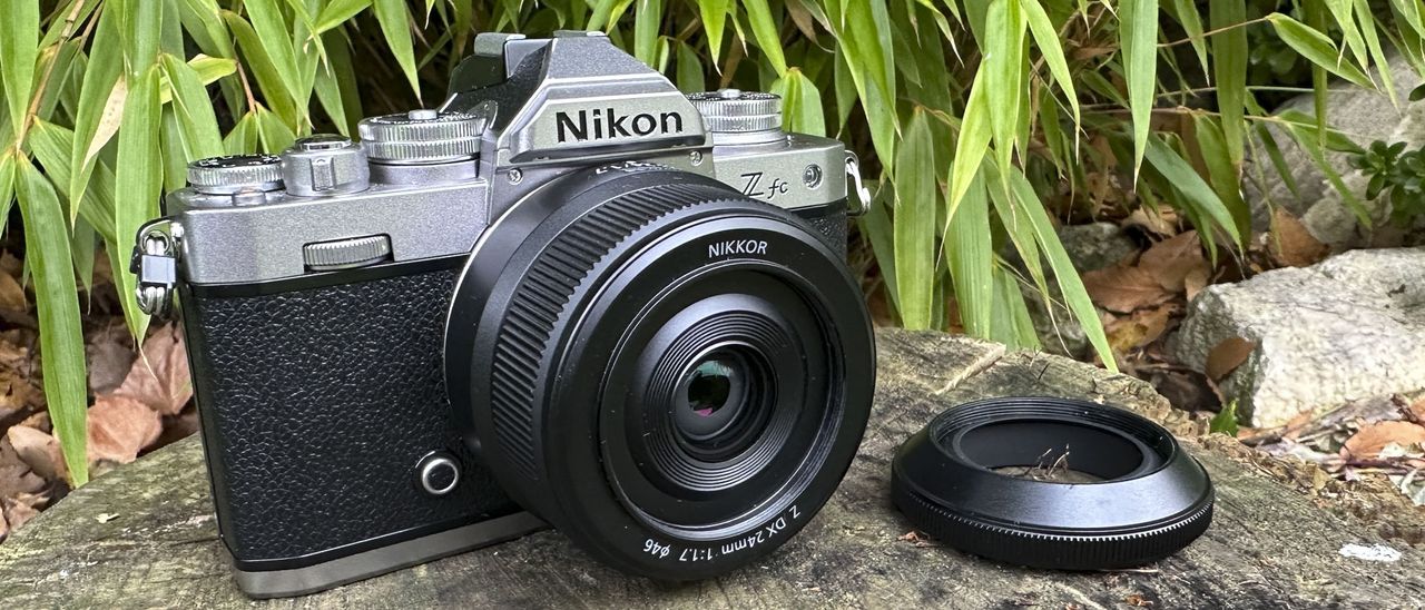The Nikkor Z DX 24mm f/1.7 lens mounted on a Nikon Z fc camera sitting on top of a log with the lens hood to one side