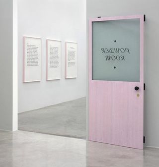 View of Matthew Brannon's show at the Casey Kaplan gallery featuring a room with light coloured walls and flooring and a pink door that says 'Powder Room'. There is a partial view of another room through the open door that has three pieces of framed text wall art on display