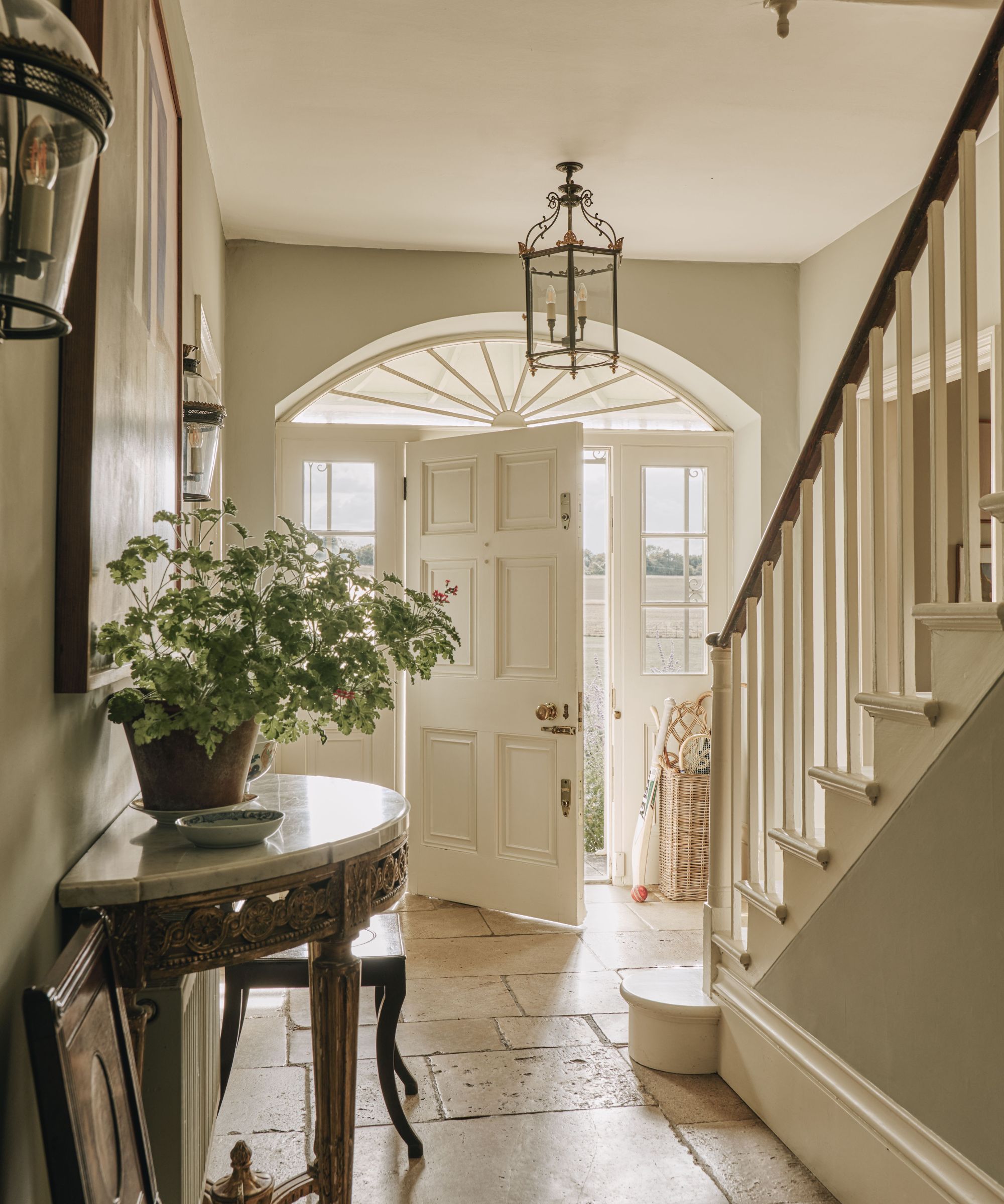Bright entryway featuring an open white front door, stone tile floor, potted green plant on a marble console table, and a wooden staircase