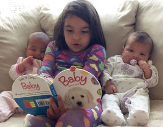 Toddler reading from teddy bear book to baby siblings on sofa