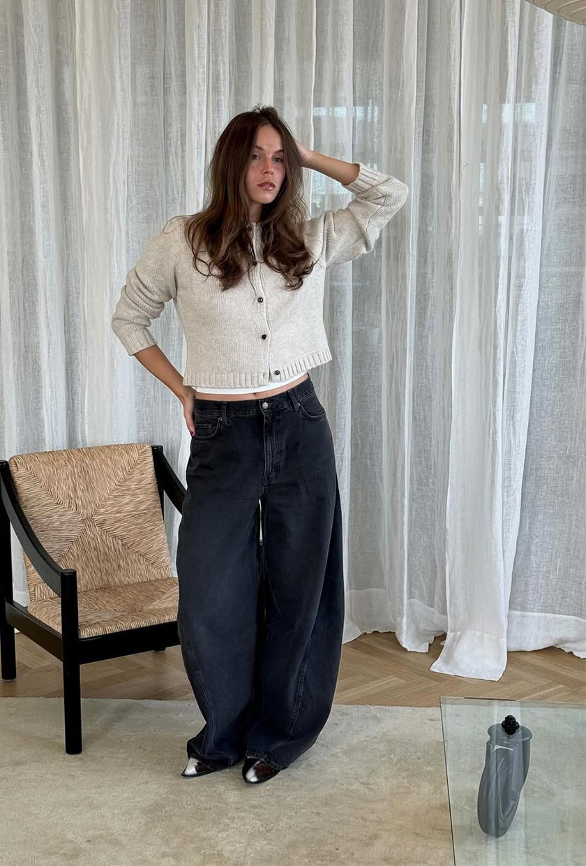 A woman standing inside her living room wearing a tan cardigan over a white T-shirt, baggy black barrel-leg jeans, and pointed cow-hair heels.