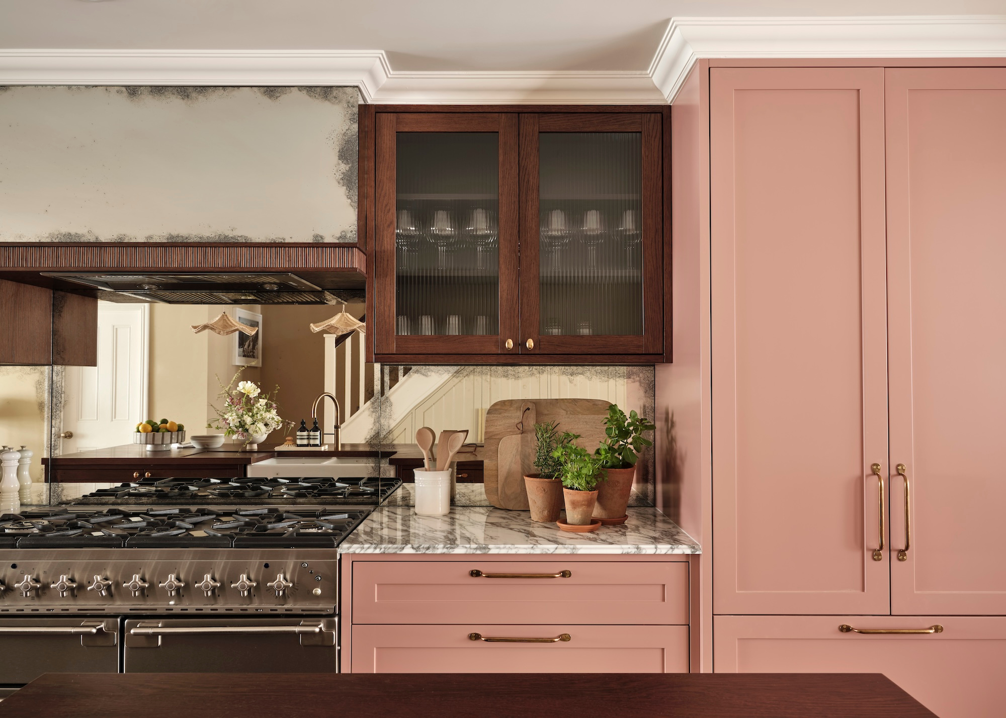A kitchen with a range cooker, dusty pink kitchen cabinets contrasted with dark wood glass fronted cabinets with a mirrored backsplash showing the stairs behind