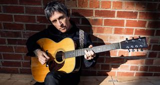 Johnny Marr sits against a brick wall and plays his new Martin M7 seven-string acoustic guitar