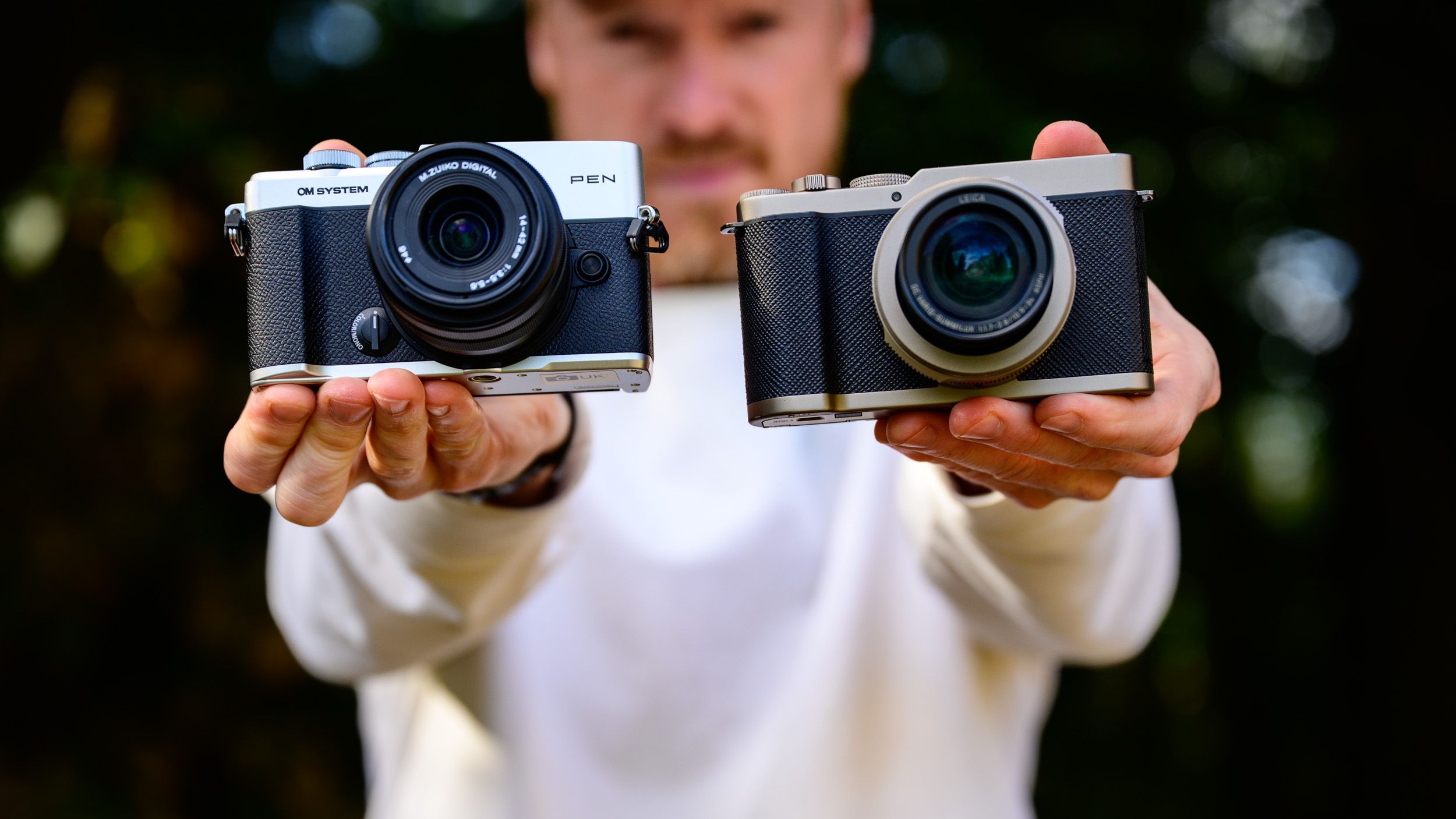 OM System PEN vs Panasonic Lumix L10: two micro four thirds cameras side by side in tech journalist Tim Colemans hands, dappled tree light background