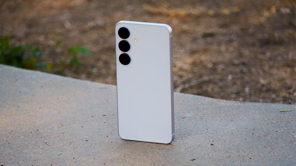 Samsung Galaxy S25 smartphone standing upright on a flat, concrete surface outdoors with three large, circular camera lenses arranged vertically near the top left corner.