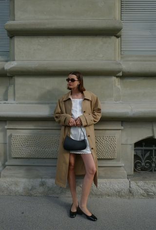 a photo of a summer rainy day outfit with a woman wearing a tan trench coat over a white minidress with black ballet flats and a black shoulder bag