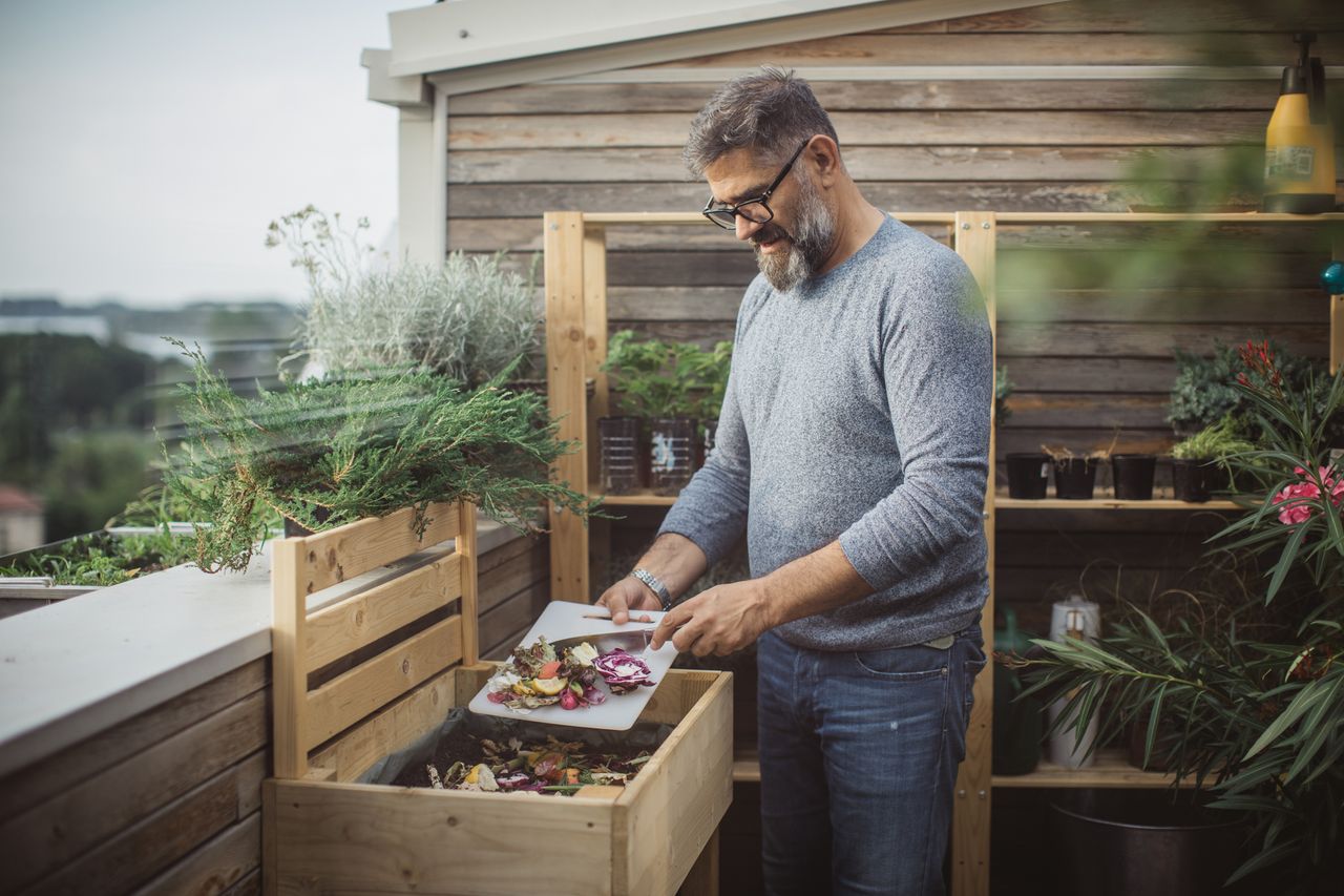 A man in glasses scrapes food scraps off a cutting board into a wooden compost bin