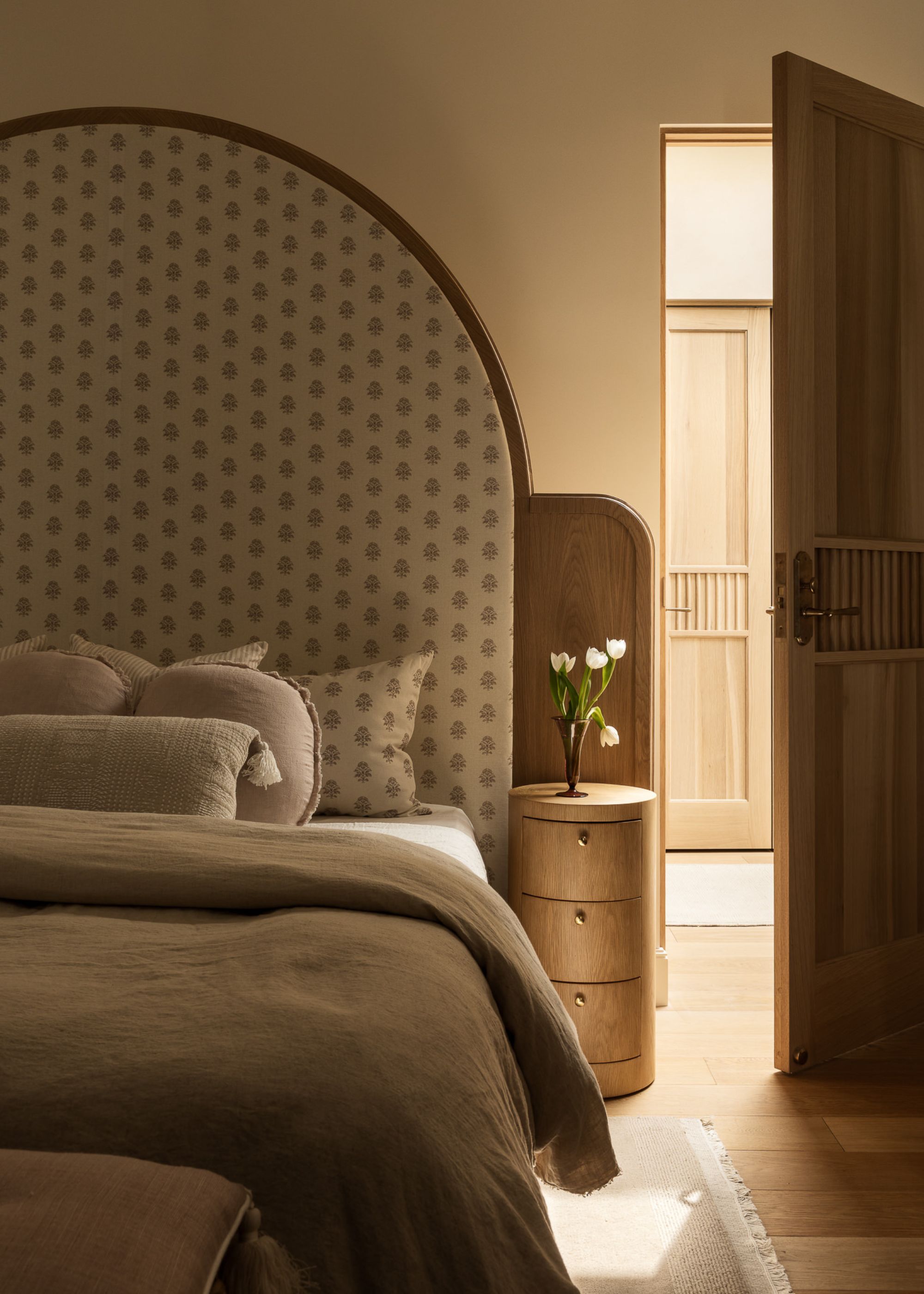 A modern neutral bedroom with a large curved headboard, a curved wooden bedside table and a wooden door with ribbed detailing in the center
