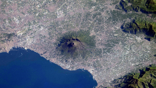 A large volcano stands out on the coastline nearby a body of water, as seen from above. 