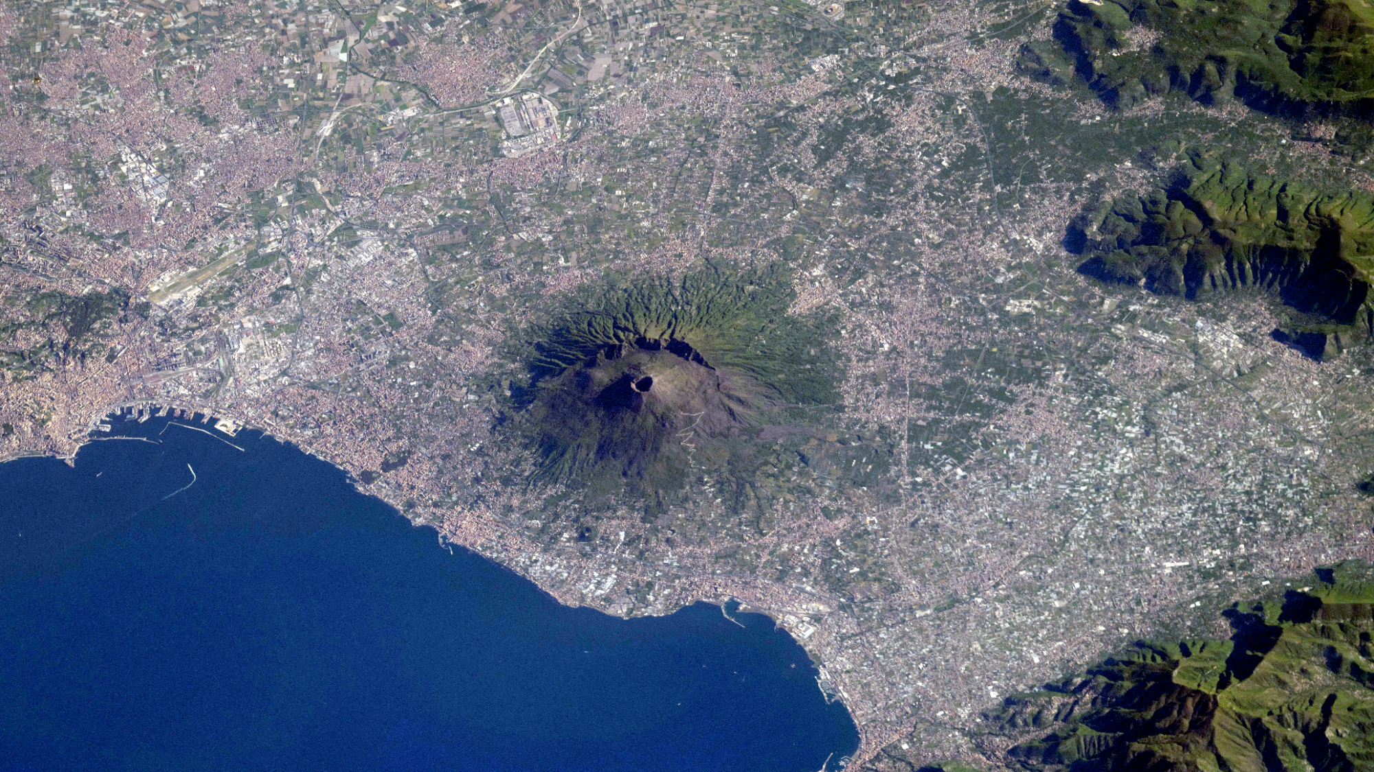 A large volcano stands out on the coastline nearby a body of water, as seen from above.