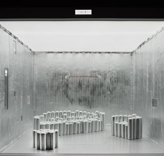 An Elevator room with padded silver walls, metal silver flooring and grouped silver slim cylinder tubes in different heights