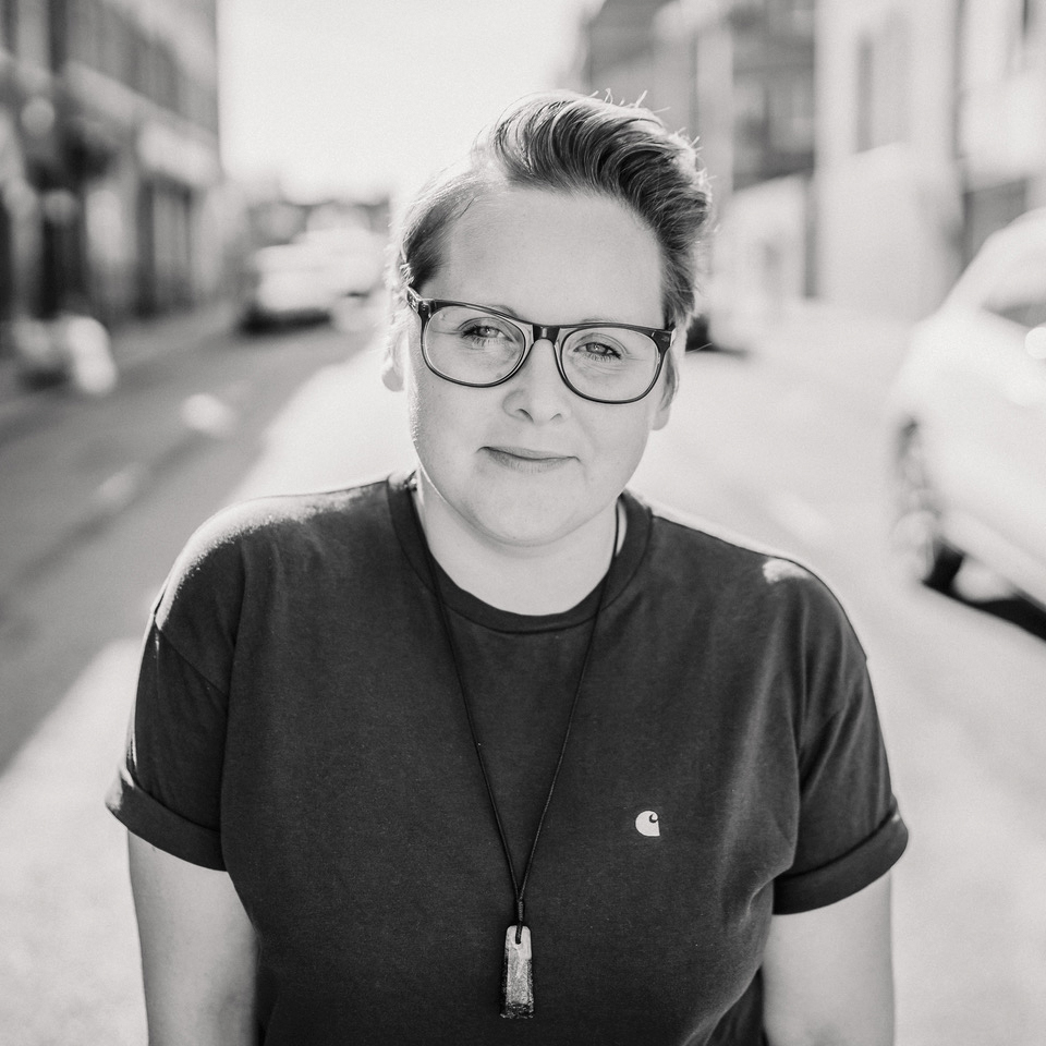 Black and white photo of a person with glasses, short hair, and a casual t-shirt, standing on a sunlit street. They appear relaxed and confident