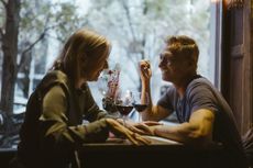 An older couple laughs together in a restaurant. They are framed by a window and drinking wine.