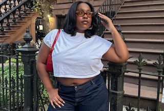 Editor Aniyah Morinia wears white t-shirt, jeans, red handbag, and tinted eyeglasses while standing in front of New York City home.