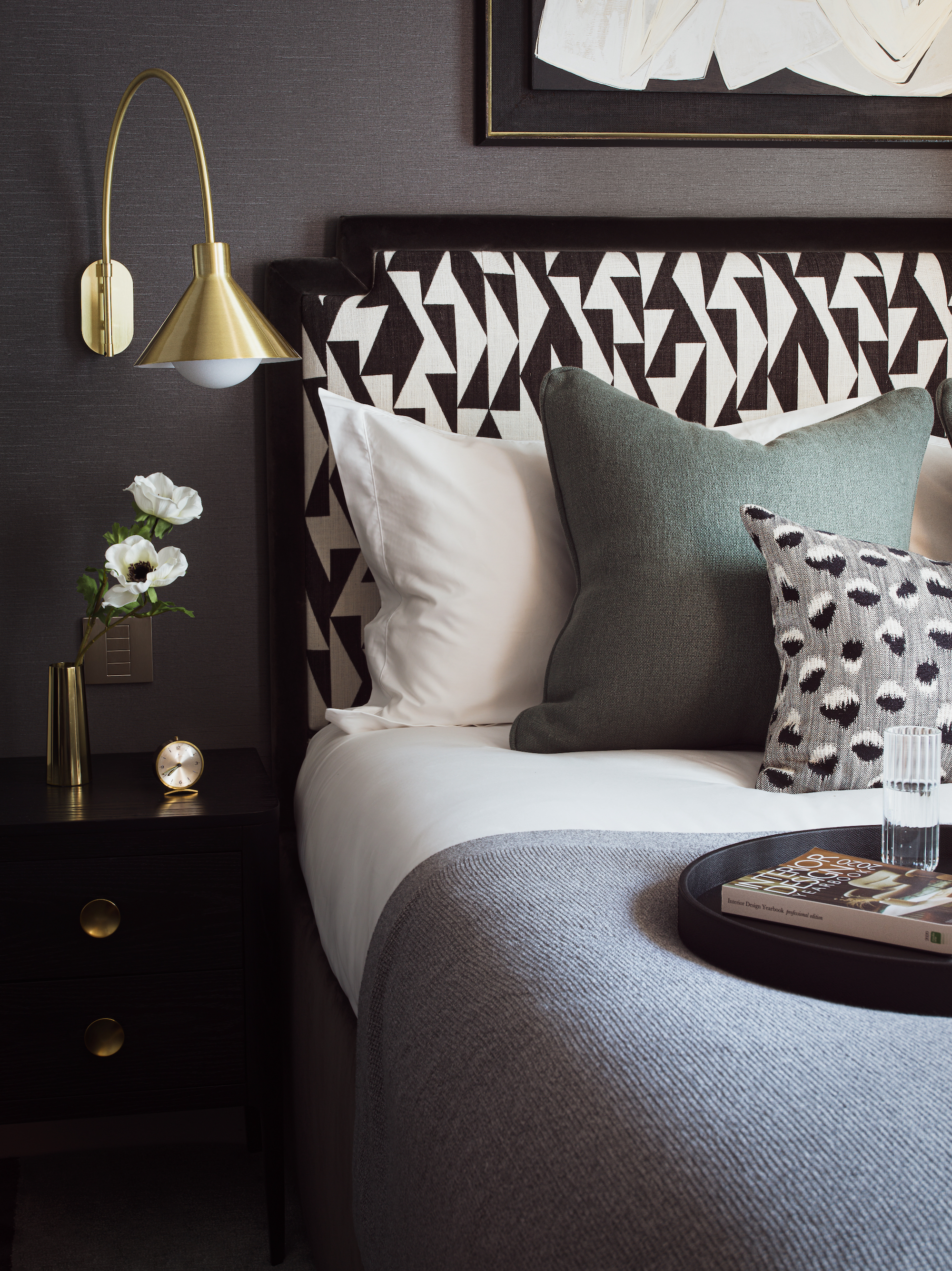 dormitorio en blanco y negro con cabecero estampado, cojines grises y blancos, cabecera negra, lámpara de pared dorada, obras de arte