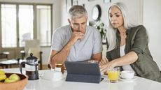 An older couple look serious as they look at a tablet together over breakfast.