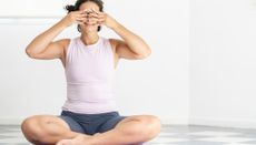 Woman sits cross legged with her fingers on her face, covering her eyes. She is wearing grey shorts and a white vest top.