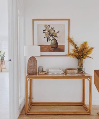 A wood and glass console table with yellow flowers, books and lamp, with wall art hanging above it depicting a lavender filled jug