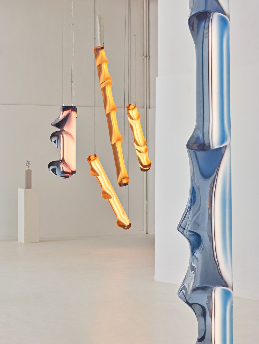 Five sculptural neon lights pending from the ceiling of an empty gallery, in pink, orange, and blue nuances, with a contorted silhouette.