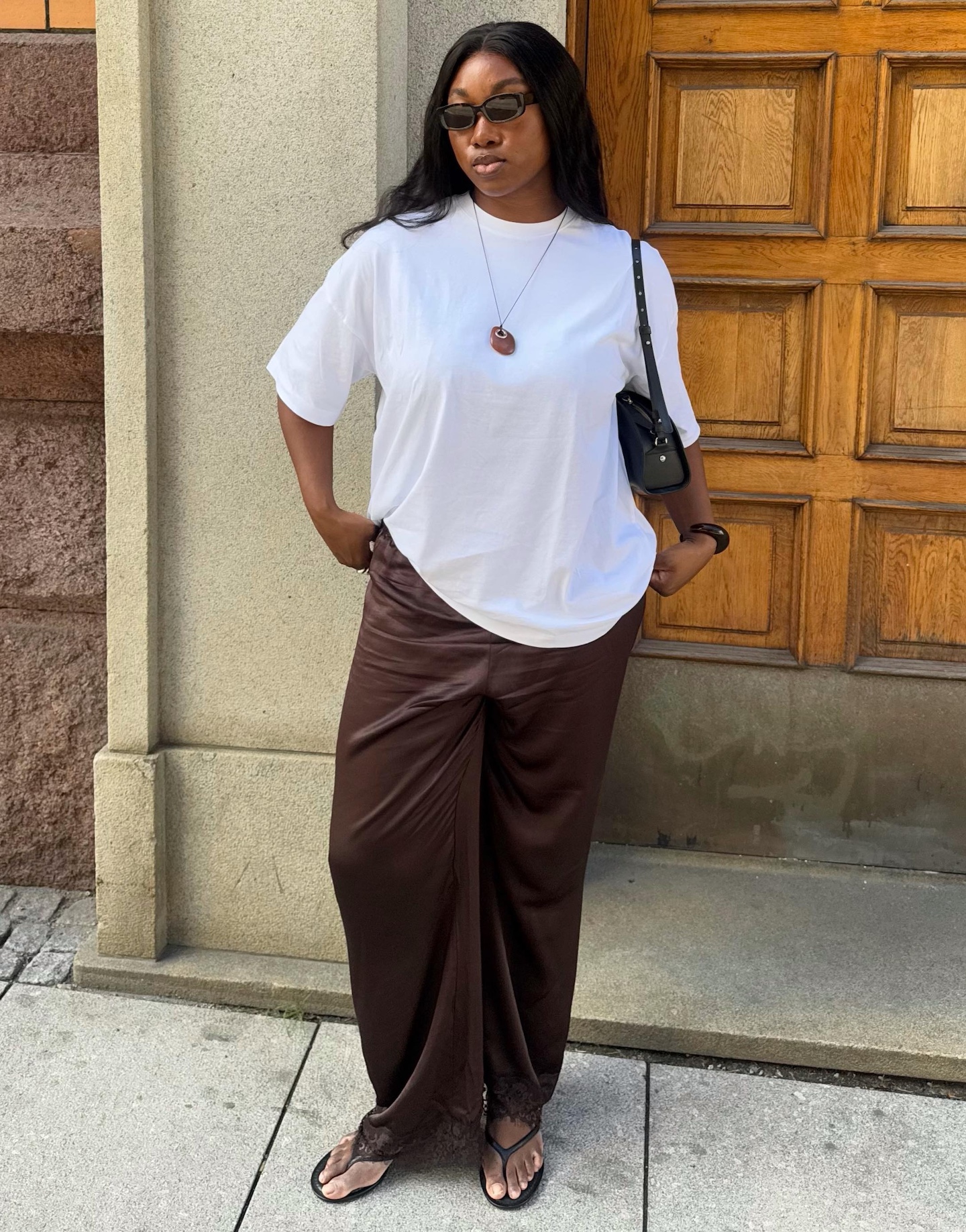 Norwegian style influencer Nnenna Echem poses in an oversize white T-shirt, a red pendant necklace, black shoulder bag, brown lace-trim satin pants, and black flip-flop sandals