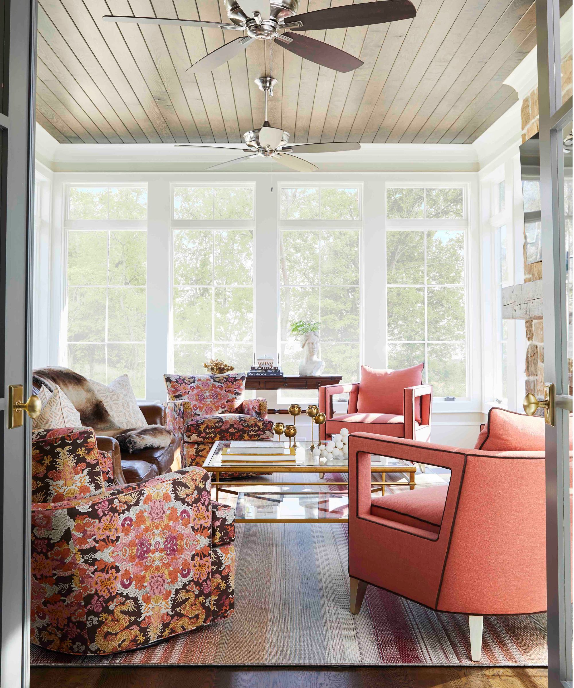 Sunroom with dark wood panelled ceiling, two ceiling fans, wrap-around floor-to-ceiling windows, white frames, a brick fireplace, and coral and floral armchairs around a glass coffee table.