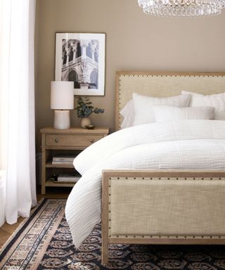 White bedding on a beige bed against beige walls; to the left, a white oak nightstand, topped with a vase and a lamp.