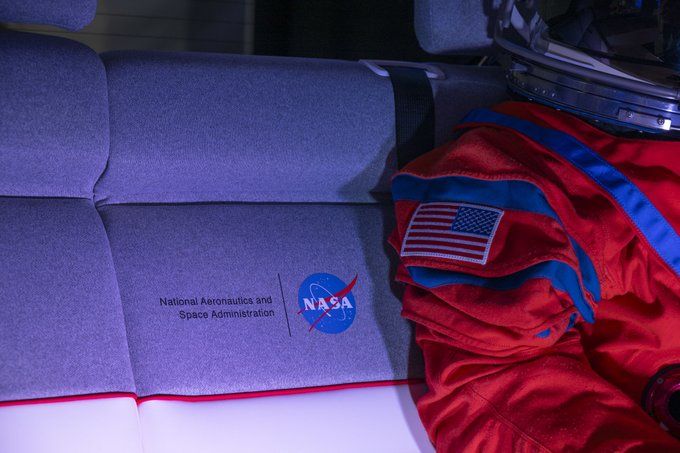 A closeup shows NASA's logo on the middle of the back seat. The seat is gray on the upper portion and white on the lower, with red piping dividing the two. The orange arm of Campos is in view.