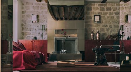 A frontal view of a chrome-clad fireplace, surrounded by lived-in brick walls, mid-century furniture, plenty of elegant collectibles, a red, draped couch, and a brown rug over white and black tiling.