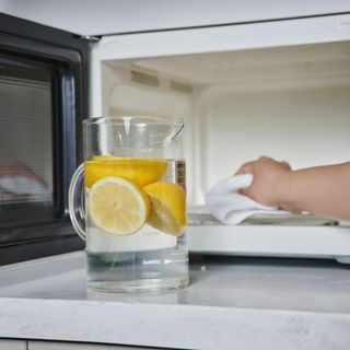 jug of water with lemons in front of a microwave and a woman's hand using a cloth to clean the microwave