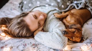 A woman with blonde-brown hair takes a nap with her sausage dog on Christmas Day, surrounded by fairy lights