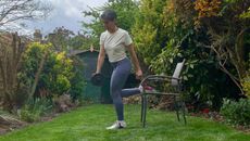 Fitness editor Yanar Alkayat performs a rear foot elevated single RDL in her yard. Her right foot rests on a chair while her left leg support her wait. She holds a dumbbell in her arm close to her thighs and her torso is slightly twisted to the right. Behind her we see bushes and a shed.